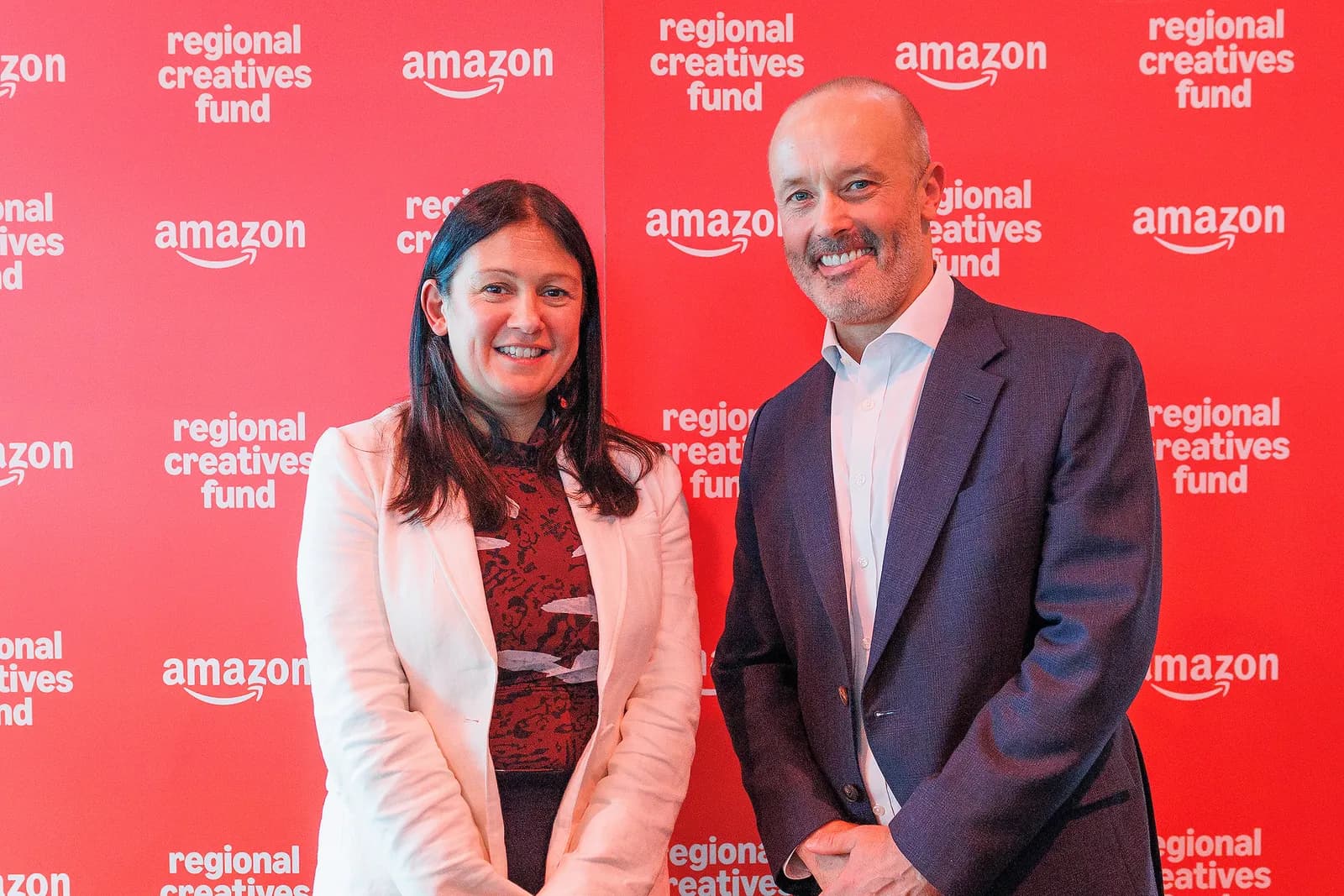 A woman and a man are smiling and standing in front of a red wall with the words "Amazon" and "regional creatives fund" repeated multiple times.