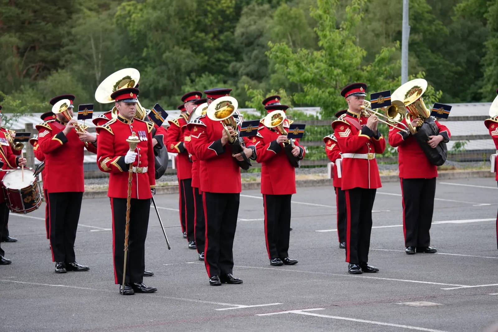 A marching band in red and black uniforms stands on an asphalt ground with trees in the background, playing brass instruments and drums.