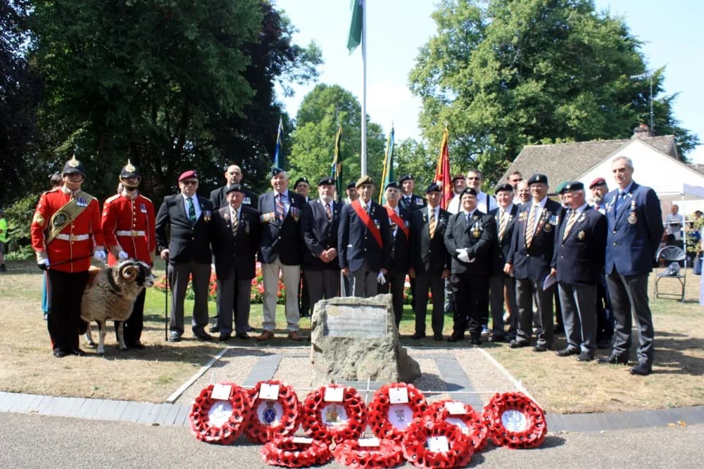 A group of uniformed men, some in ceremonial red coats and others in suits, stand around a war memorial with a sheep next to the man in the far left red coat, with several poppy wreaths laid in front of the memorial.