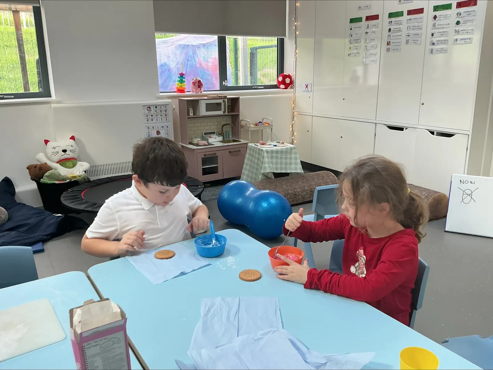 Two young children are seated at separate blue tables, each with a biscuit and bowls of colored icing, appearing to be decorating them in a brightly lit room.