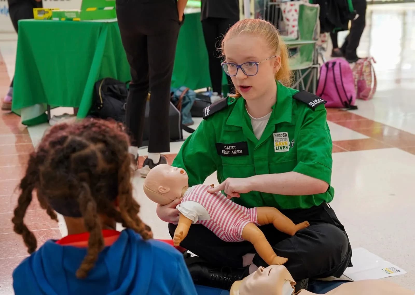 A young woman in a green uniform demonstrates CPR on a baby mannequin to a child.