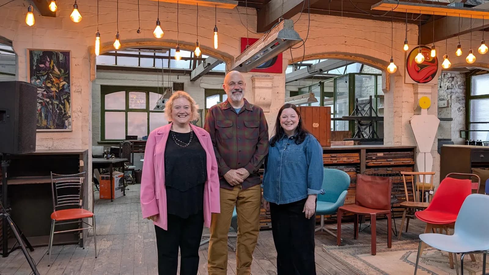 Three people stand smiling for a photo in a room with exposed brick walls, hanging lightbulbs, and various chairs.