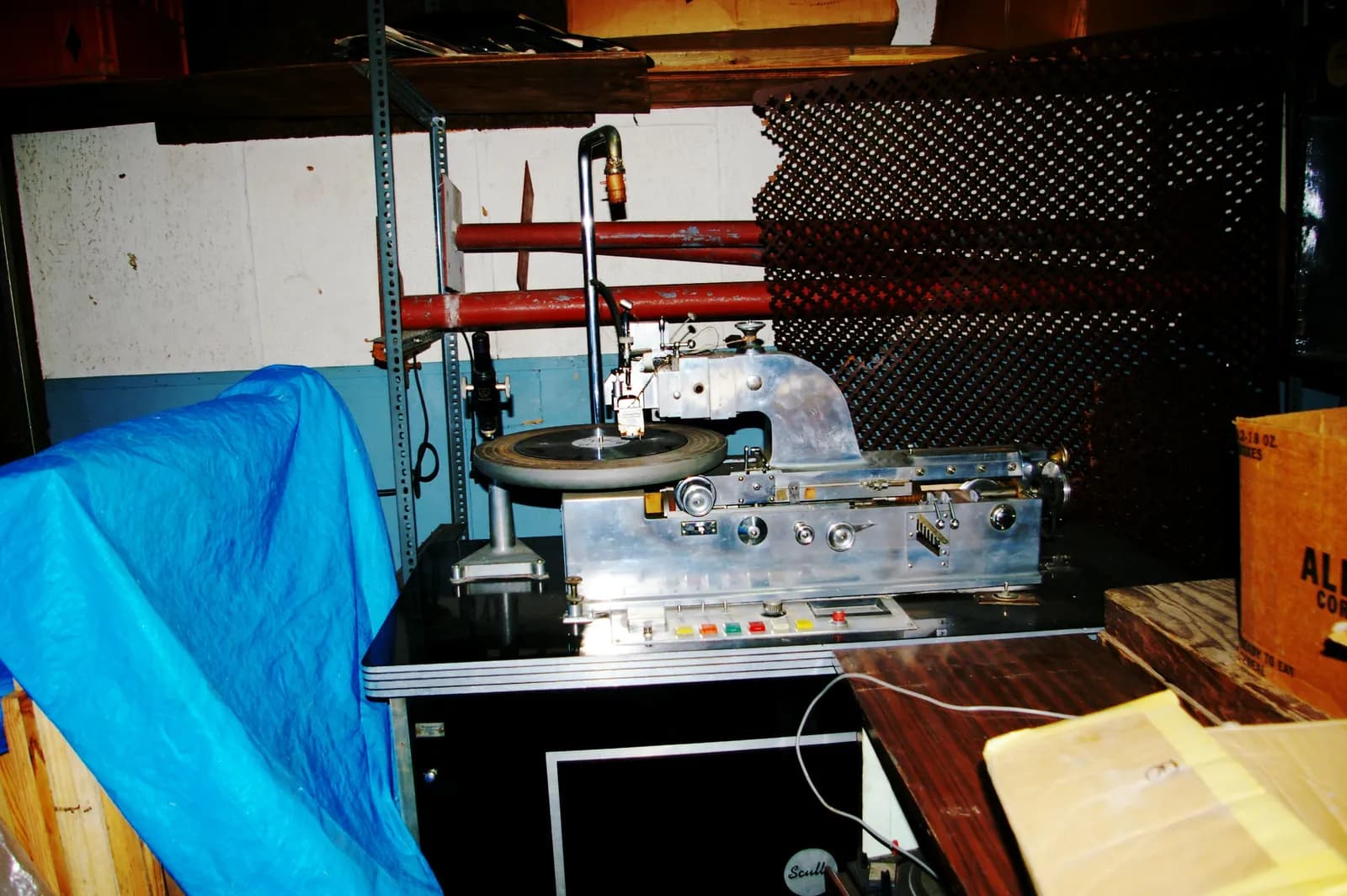 A vintage Scully record lathe, a large silver and black machine with several red pipes behind it, sits on a black table with a blue tarp and wooden crates around it in a cluttered room.