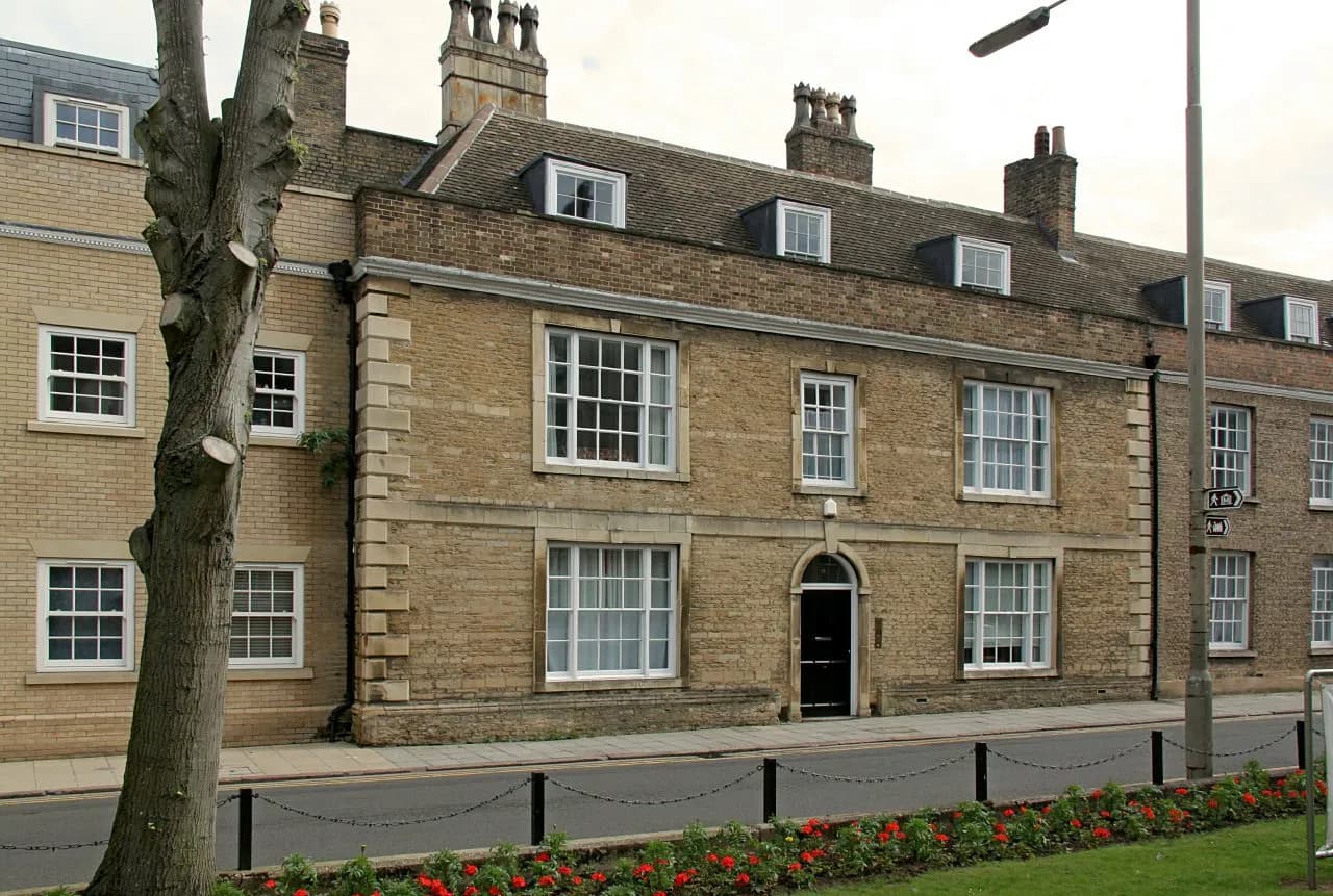 A row of historic brick buildings with white-paned windows and a central black door, alongside a street with a tree in the foreground and red flowers in a garden bed.