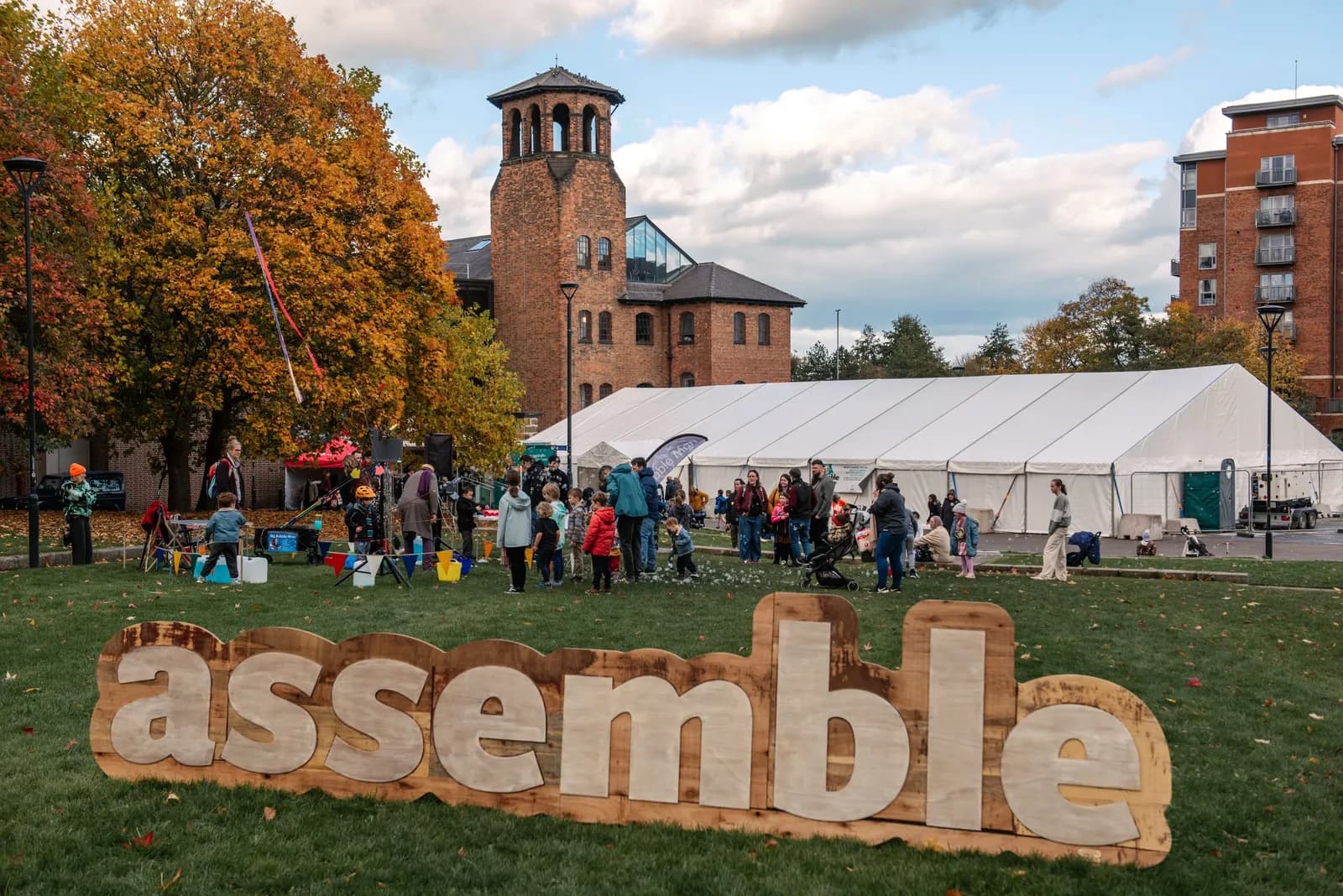 A large wooden sign reading "assemble" in white letters sits on a grassy lawn with a brick building and a white event tent in the background, surrounded by people.