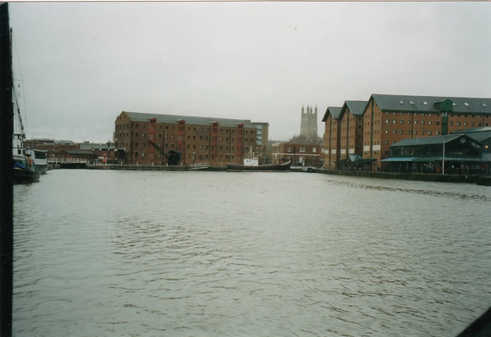 A wide shot of warehouses lining a calm body of water with a distinct church tower visible in the distance under an overcast sky.