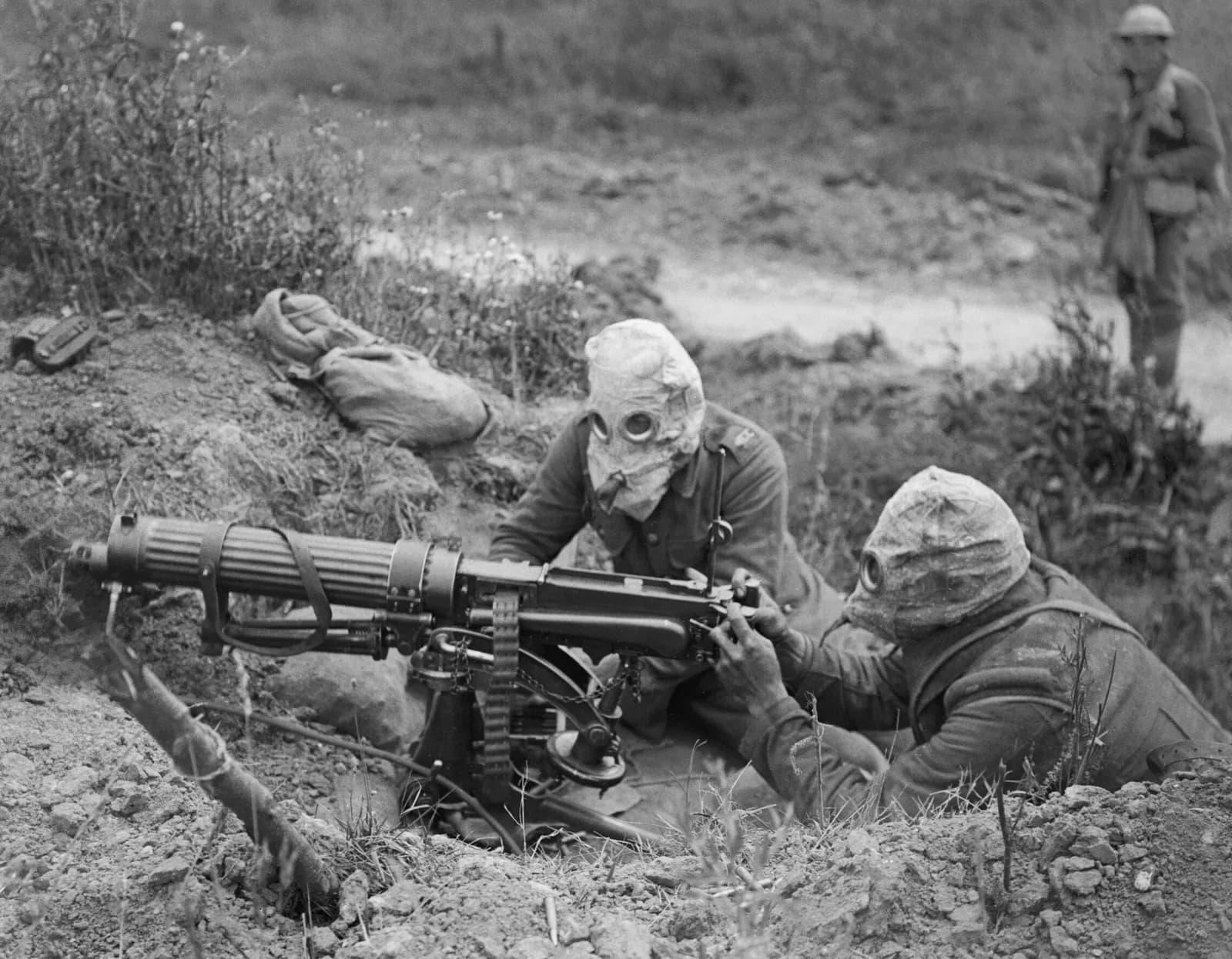 Two soldiers in gas masks operate a machine gun from a trench, with another soldier standing in the background.
