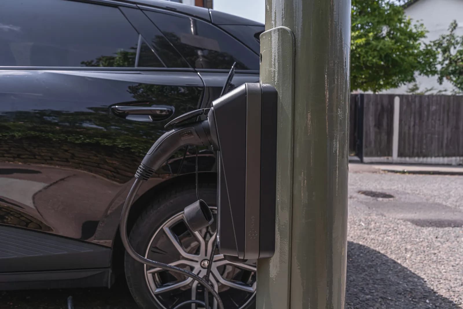 A black electric car is charging from a grey charging station mounted on a green utility pole.