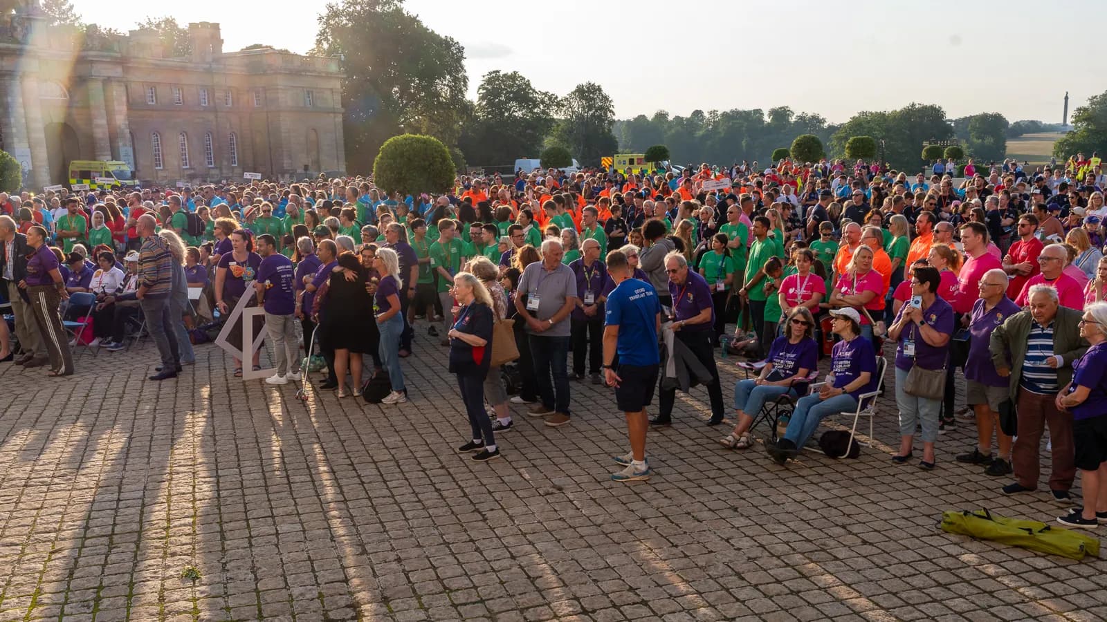 A large crowd of people, many wearing purple, green, and orange shirts, gather outdoors on a paved area with a large building and trees in the background.