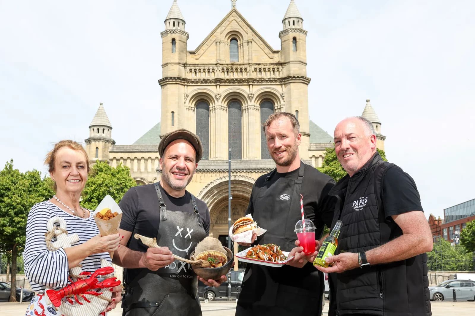 Four people, two men in black chef's aprons, a man in a black vest, and a woman in a striped shirt, holding various food and drinks in front of a large stone building with towers.