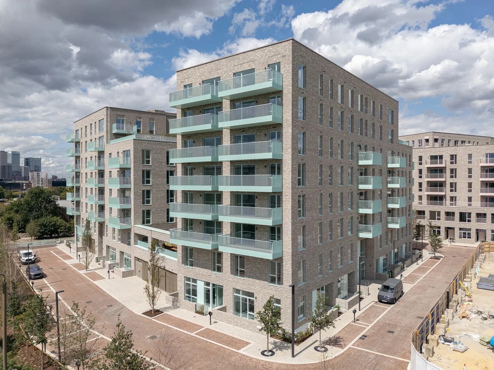A modern residential building with light brown brickwork and aqua-colored balconies stands under a partly cloudy sky.