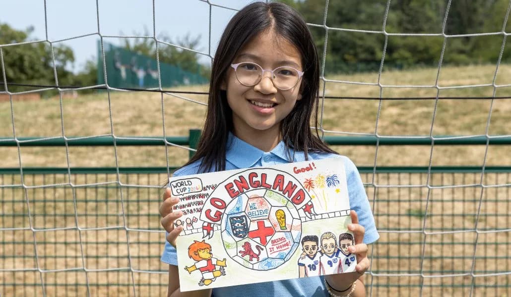 A young girl with long dark hair and glasses smiles while holding up a hand-drawn football-themed poster that says 'Go England!'.