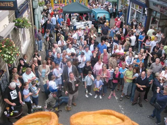 A large crowd of people of all ages fills a narrow street, with buildings lining both sides and hanging baskets with flowers visible.