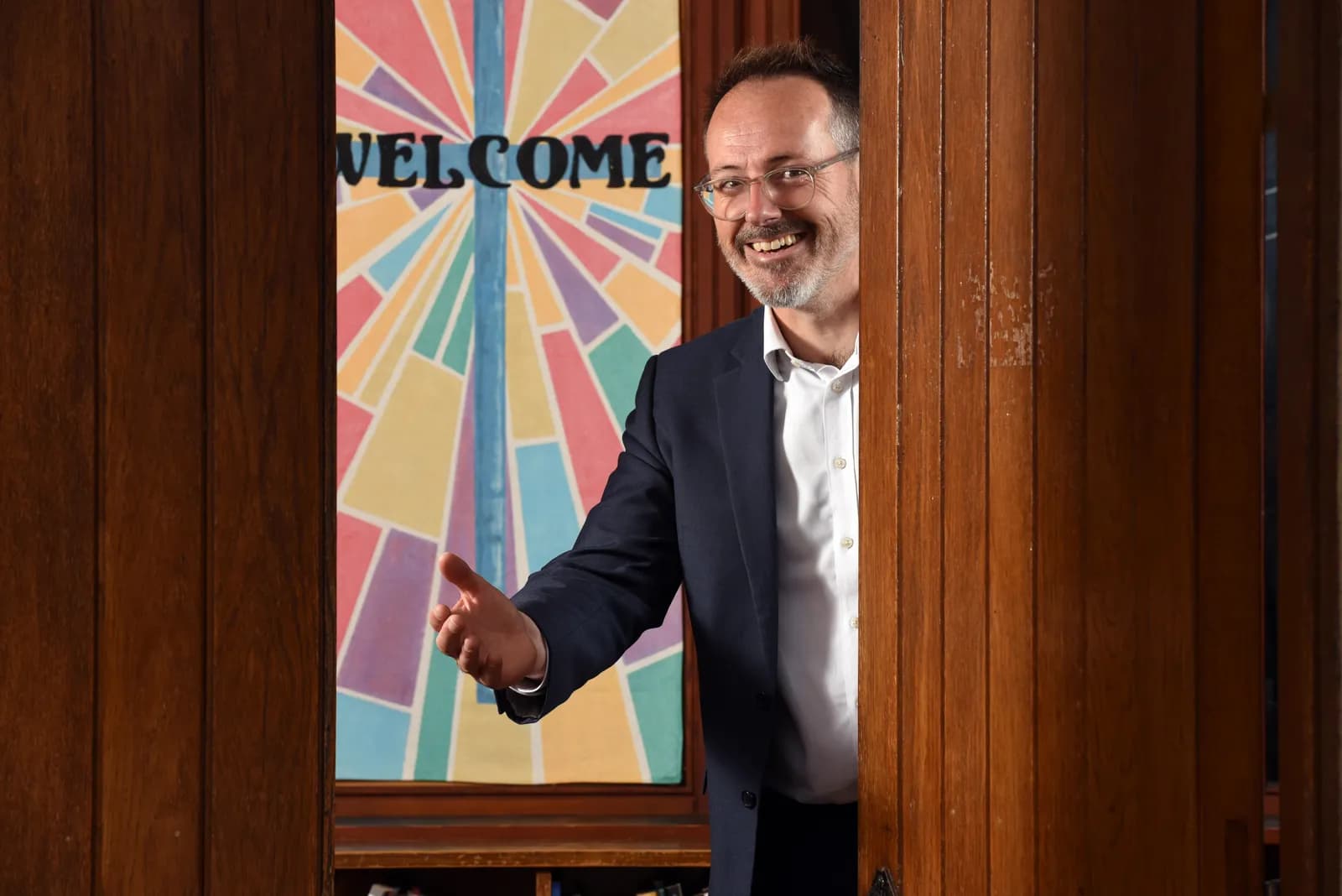A smiling man with a beard and glasses, wearing a suit jacket and white shirt, peeks from behind a wooden door with his arm extended, as if to invite someone in. Behind him is a colorful stained-glass style sign that reads "WELCOME".
