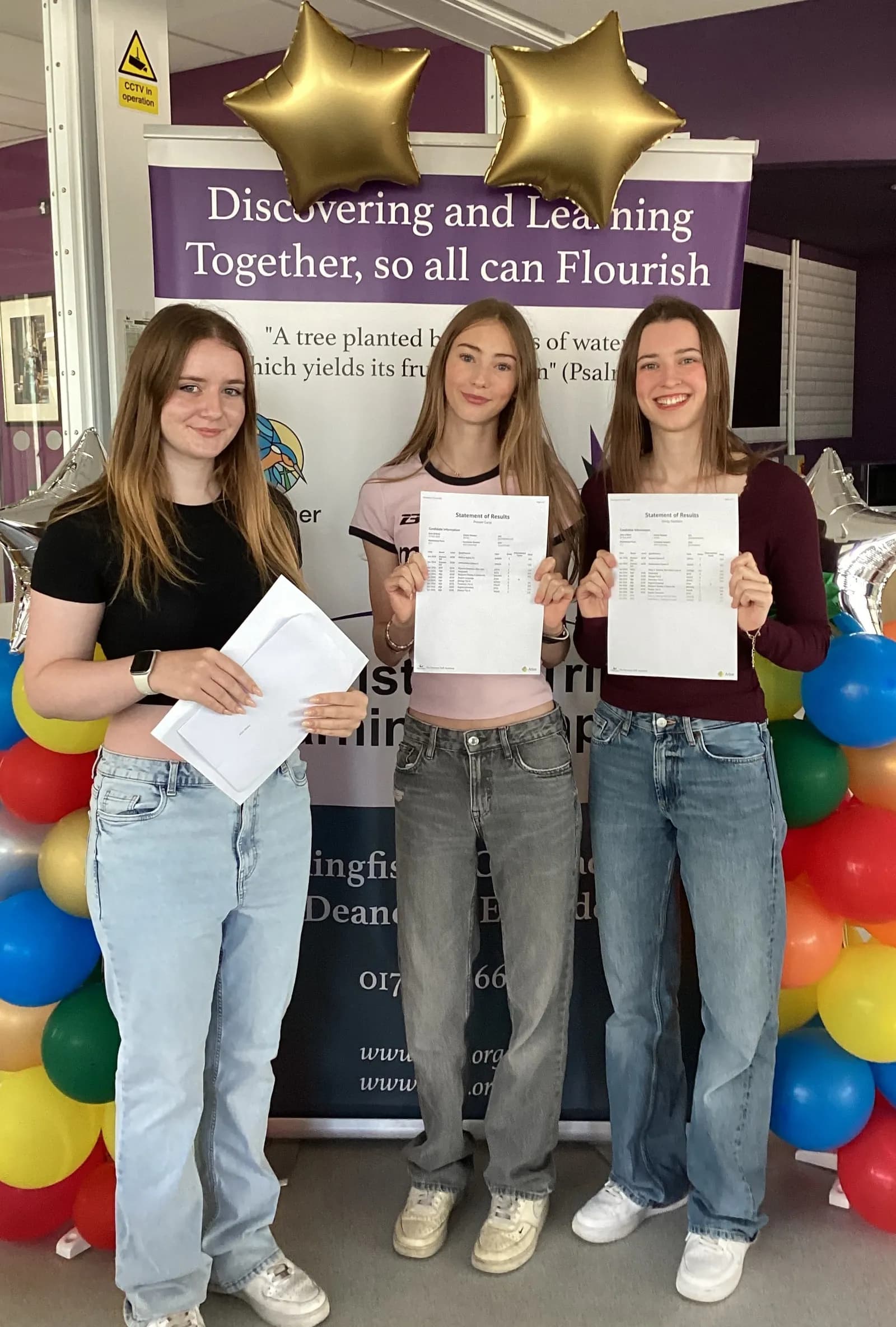 Three young women stand smiling in front of a banner and balloons, holding up papers that appear to be results or certificates.