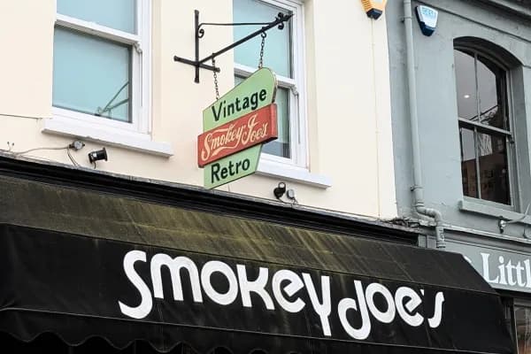 A black awning with the white lettering "Smokey Joe's" partially visible, with a hanging sign above it that reads "Vintage Smokey Joe's Retro" in green and red against a cream-colored building with white-framed windows.
