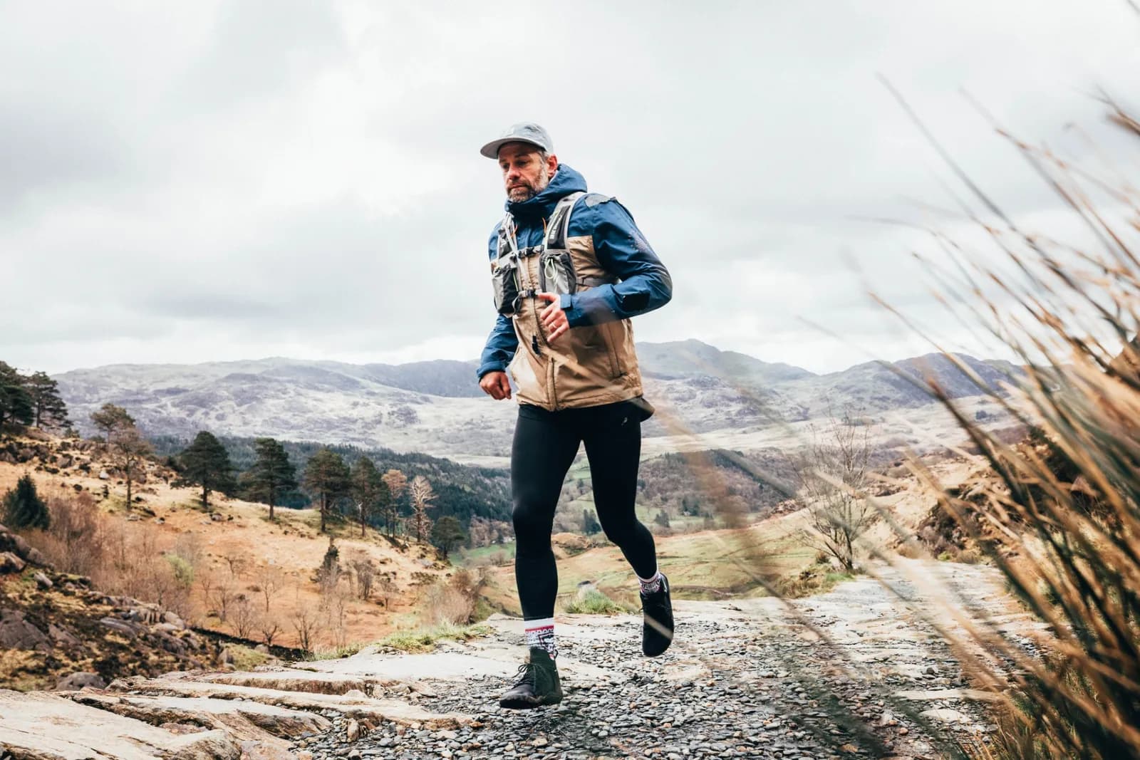 A man in athletic wear, including a cap, blue jacket, and tan vest, is trail running on a rocky path through a mountainous and hilly landscape under a cloudy sky.