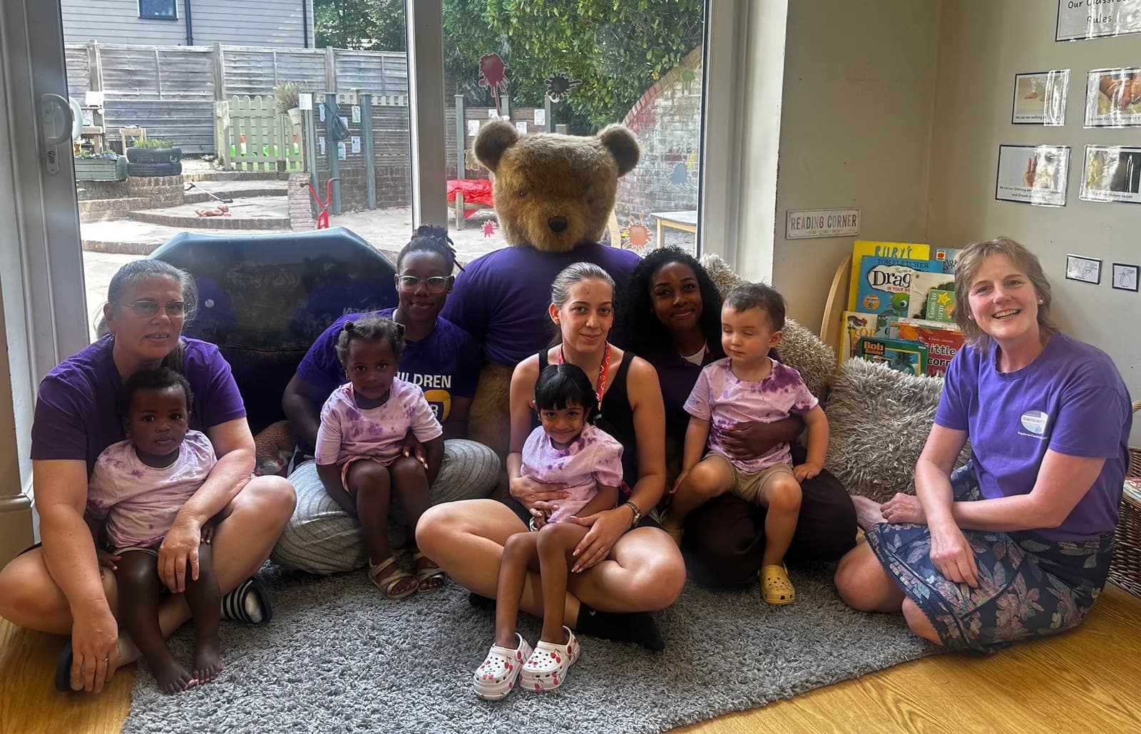 A group of adults and children are sitting on a rug in a room, with a large teddy bear behind them.