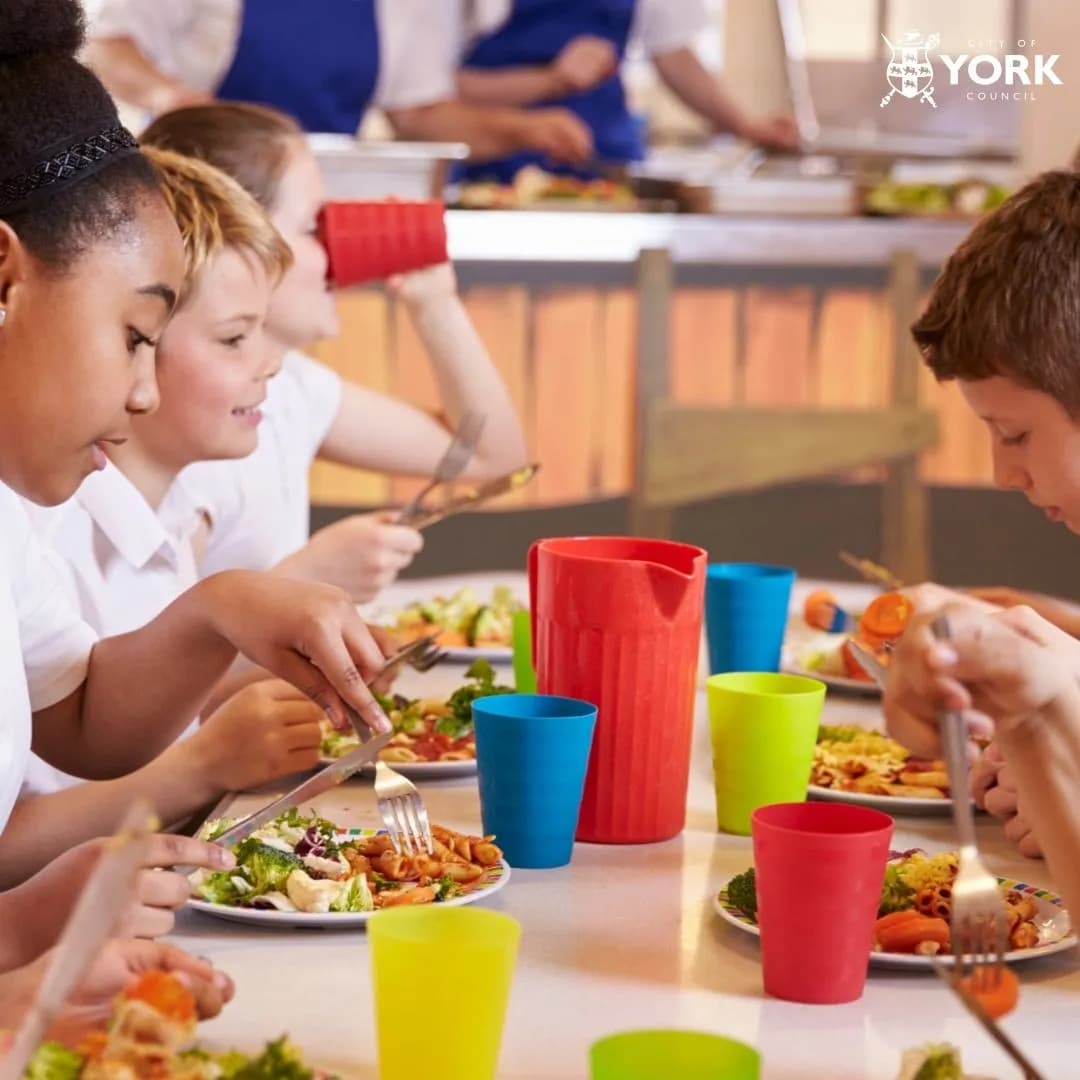 Several children in white shirts happily eating meals with forks and knives at a long table with colorful plastic cups and pitchers in a cafeteria setting.
