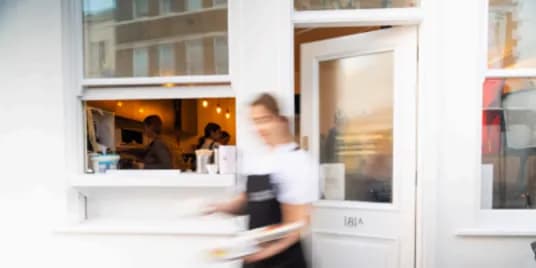 A blurred figure of a server carrying plates emerges from a cafe's takeaway window, with other staff visible inside.