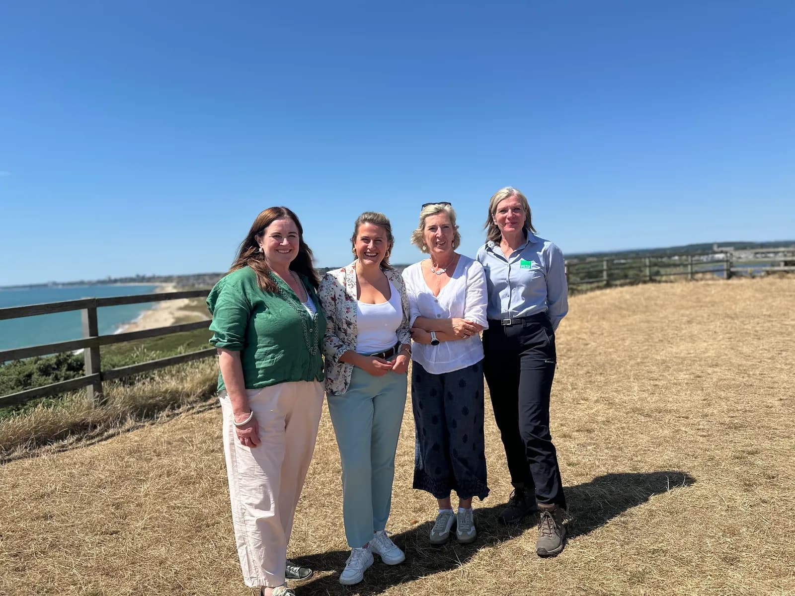 Four women standing on a grassy hill overlooking a coastline on a clear day.