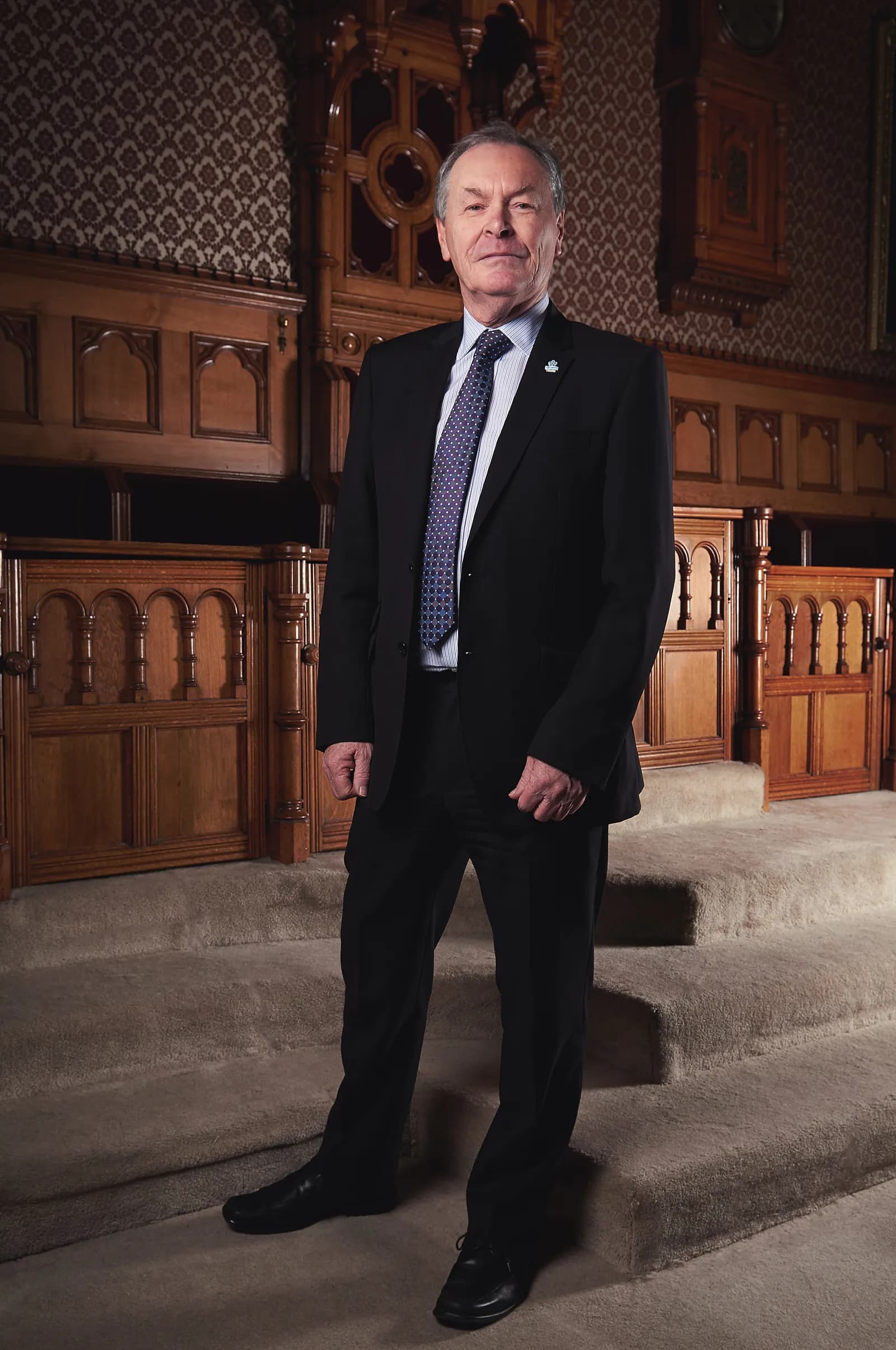 A man in a black suit and blue tie stands on carpeted steps in front of ornate wooden walls with patterned wallpaper.