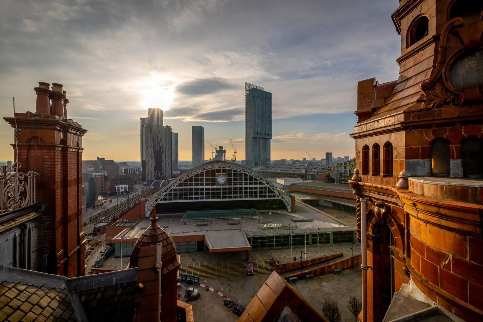 A wide shot of Manchester's city center, featuring the arched roof of Manchester Central Convention Complex, modern skyscrapers, and traditional brick buildings, all bathed in the warm light of a sunset or sunrise.