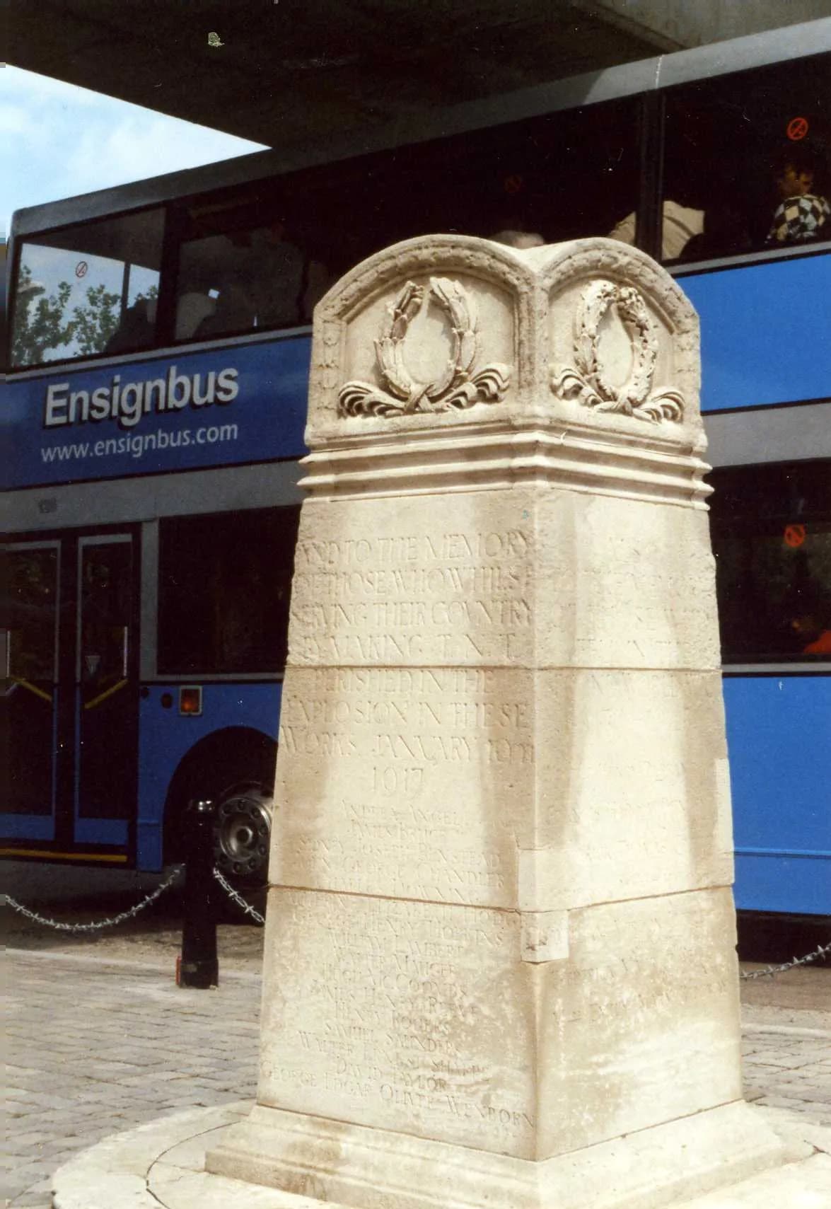 A stone monument with carved details and inscriptions stands in the foreground, with a blue double-decker Ensignbus visible in the background.