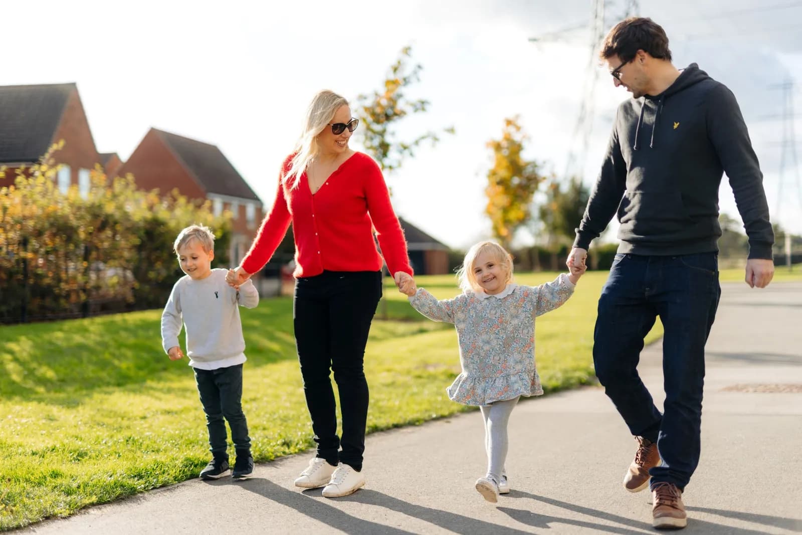 A family of four, including a mother in red, a father in grey, and two young children, walk hand-in-hand along a paved path with houses and green grass in the background on a sunny day.