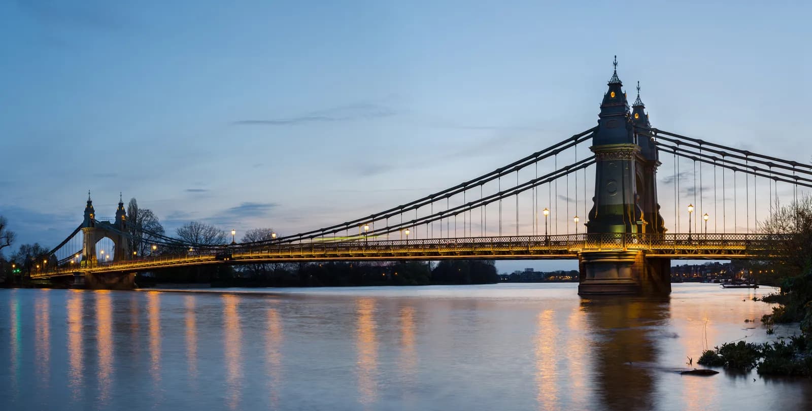 A long suspension bridge with illuminated streetlights stretches across a wide river at dusk, with its golden lights reflecting on the water.