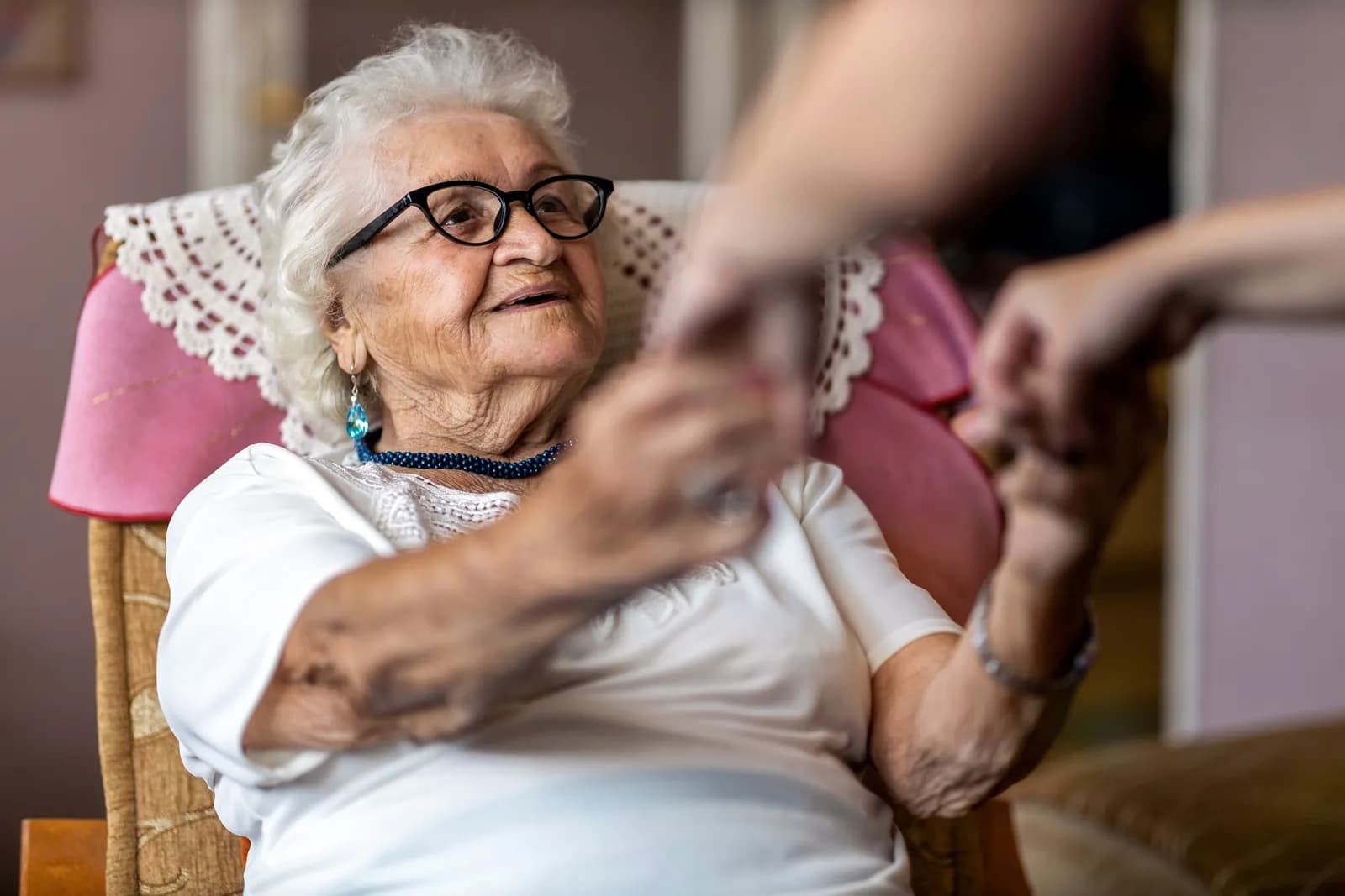 An elderly woman with white hair and glasses, wearing a white shirt and a blue beaded necklace, sitting in a patterned chair, holding hands with an unseen person.