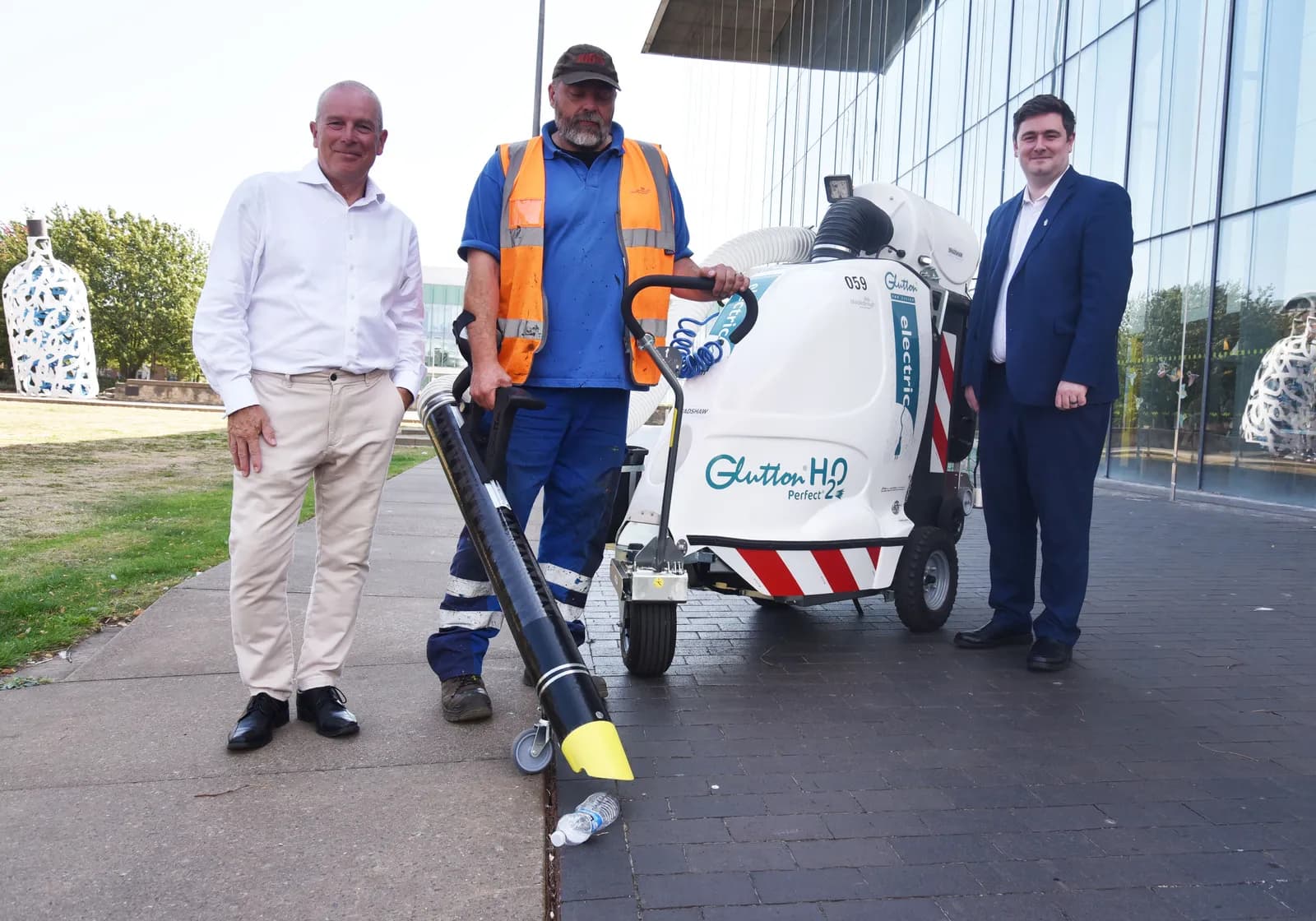 Three men stand outside a modern building with a street cleaning machine and a litter vacuum.