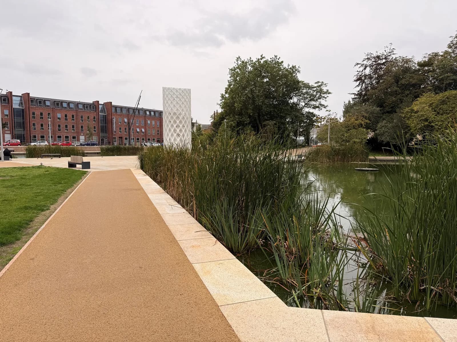 A pathway leads along a park with a modern white sculpture, a pond surrounded by tall grasses, and a red brick building in the background.