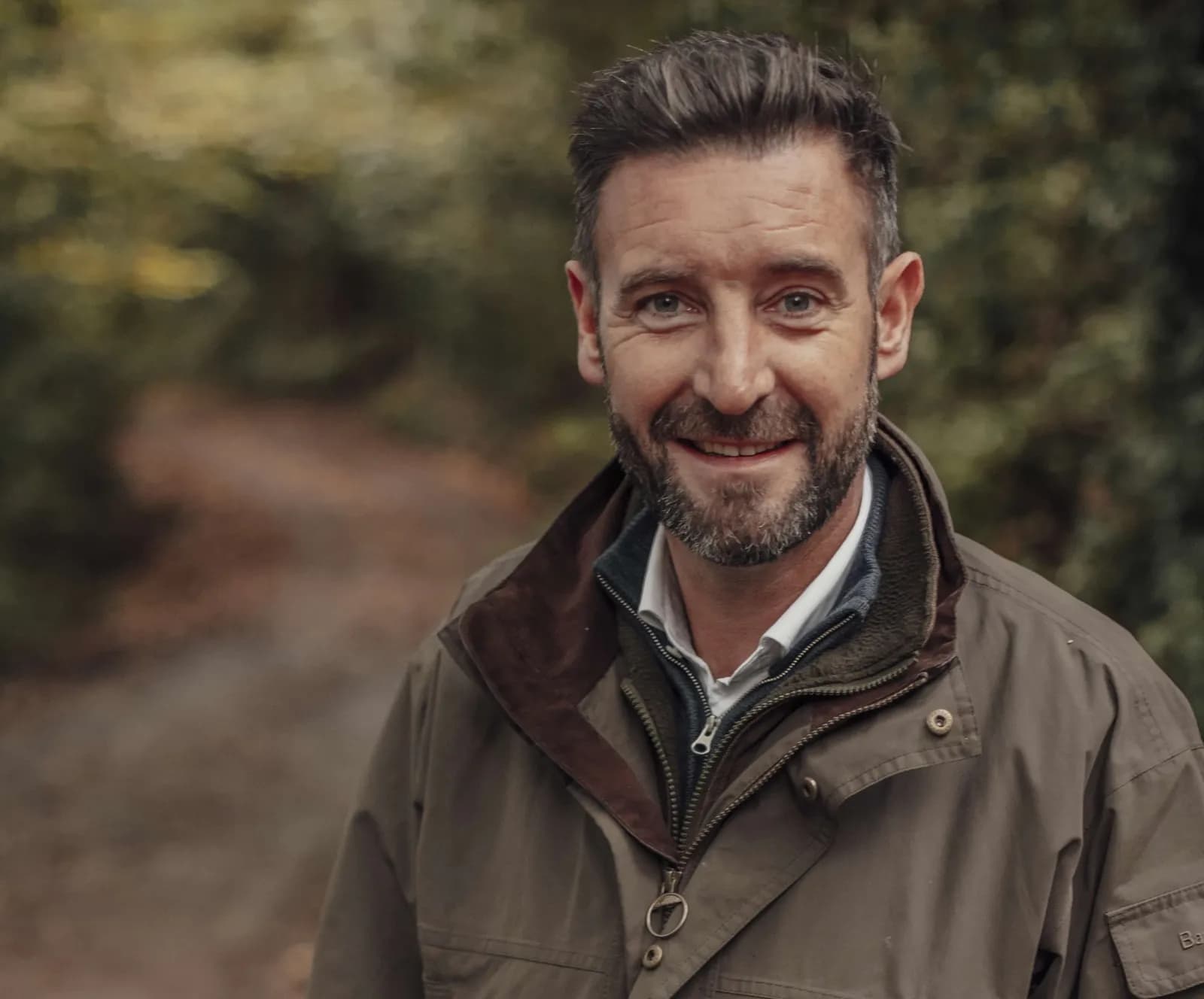 A smiling man with a beard and a short haircut is wearing a brown jacket outdoors.