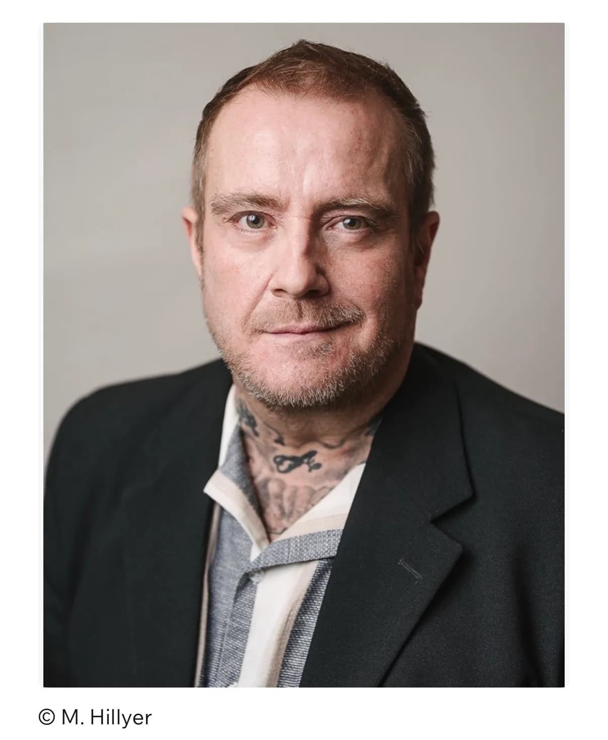 A man with light brown hair, a beard, and neck tattoos wears a dark blazer and a striped collared shirt, looking directly at the viewer.