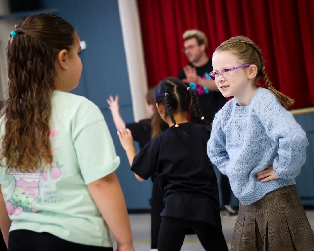 A young girl with blonde hair in pigtails and purple glasses smiles while looking to her left during a group activity, with another girl and an adult in the background.