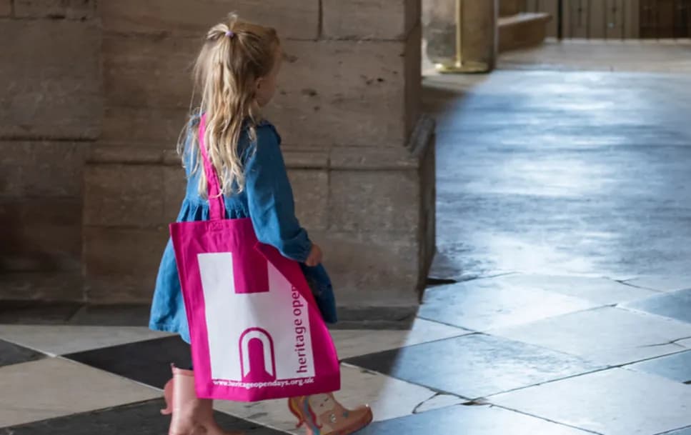 A young girl with blonde hair in a ponytail, wearing a blue dress and carrying a bright pink tote bag, walks through a hallway with stone columns and a checkered floor.