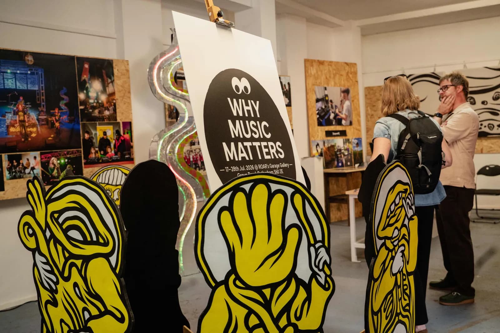 A man and a woman viewing artwork in a brightly lit gallery, with a sign that reads 'WHY MUSIC MATTERS' prominently displayed.