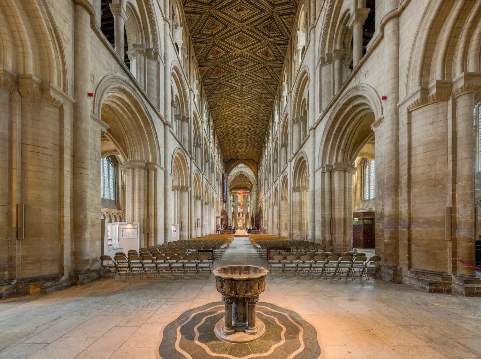 A long, ornate church interior with a patterned ceiling, rows of chairs, and a baptismal font in the foreground.