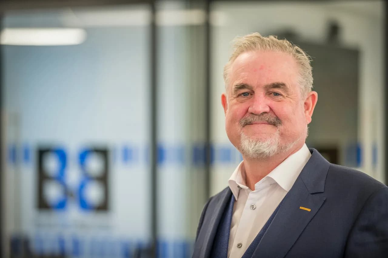 A smiling man with a gray beard and hair, wearing a white shirt and blue suit jacket, looks directly at the viewer.