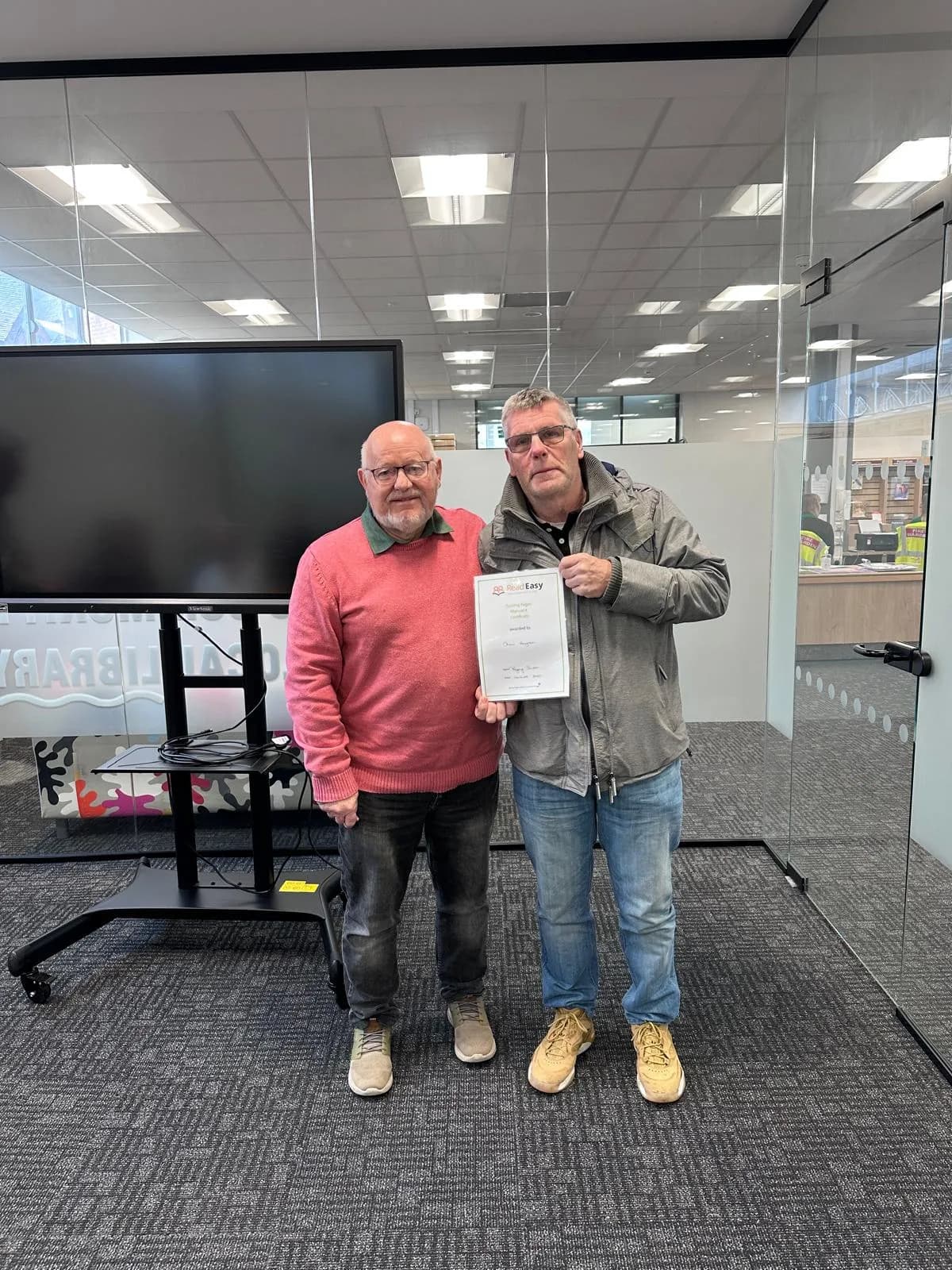 Two men standing next to each other, one in a pink sweater and the other in a gray jacket, holding a certificate.