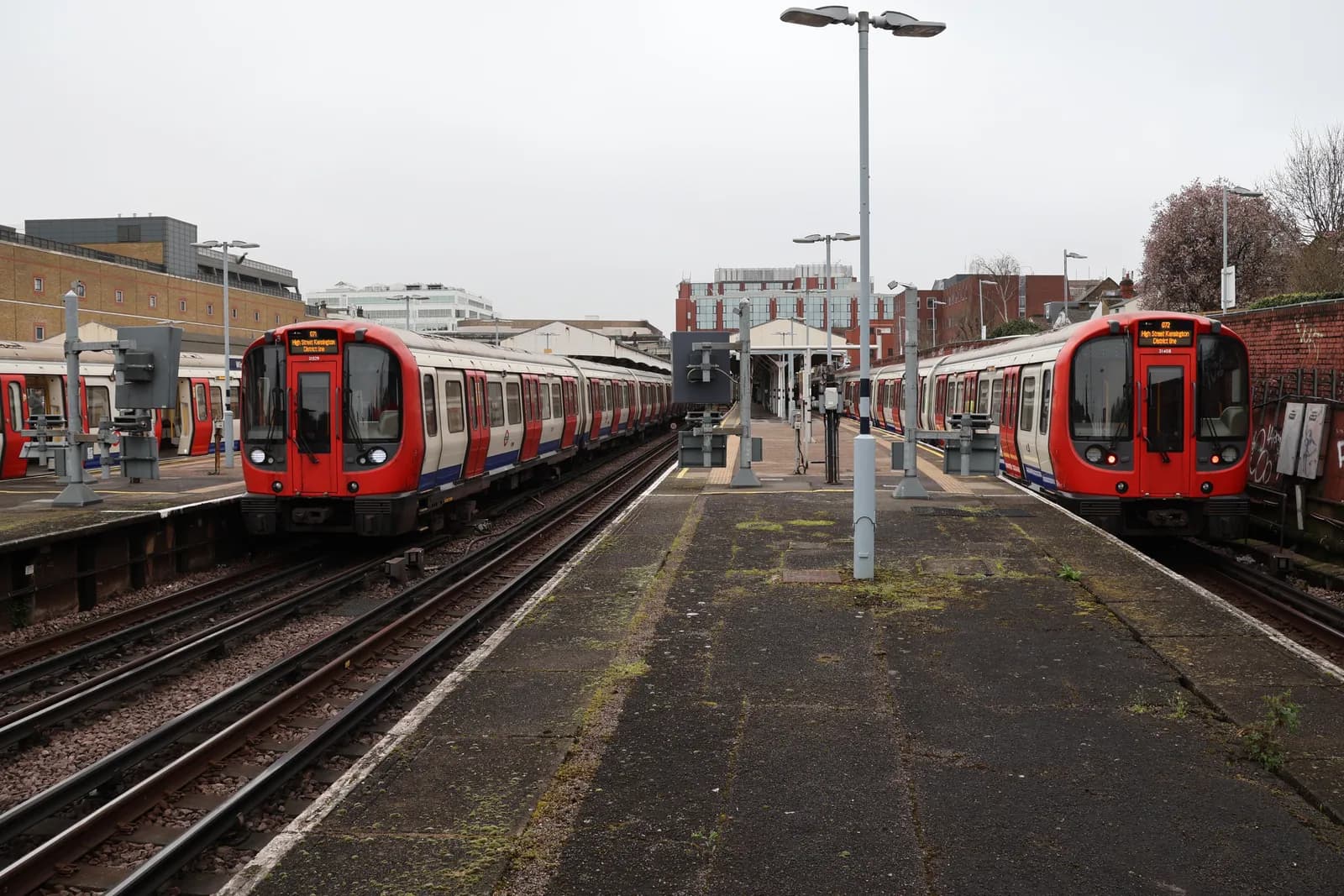 Two red Tube trains are in a station, with one pulling in on the left and another waiting on the right, under a gray sky.
