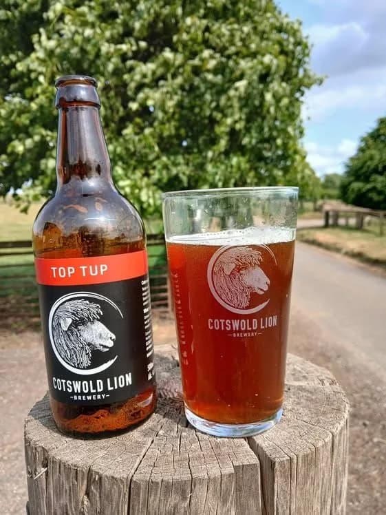 A bottle of "Top Tup" beer and a glass of beer from Cotswold Lion Brewery sit on a tree stump in front of lush green trees and a dirt path.