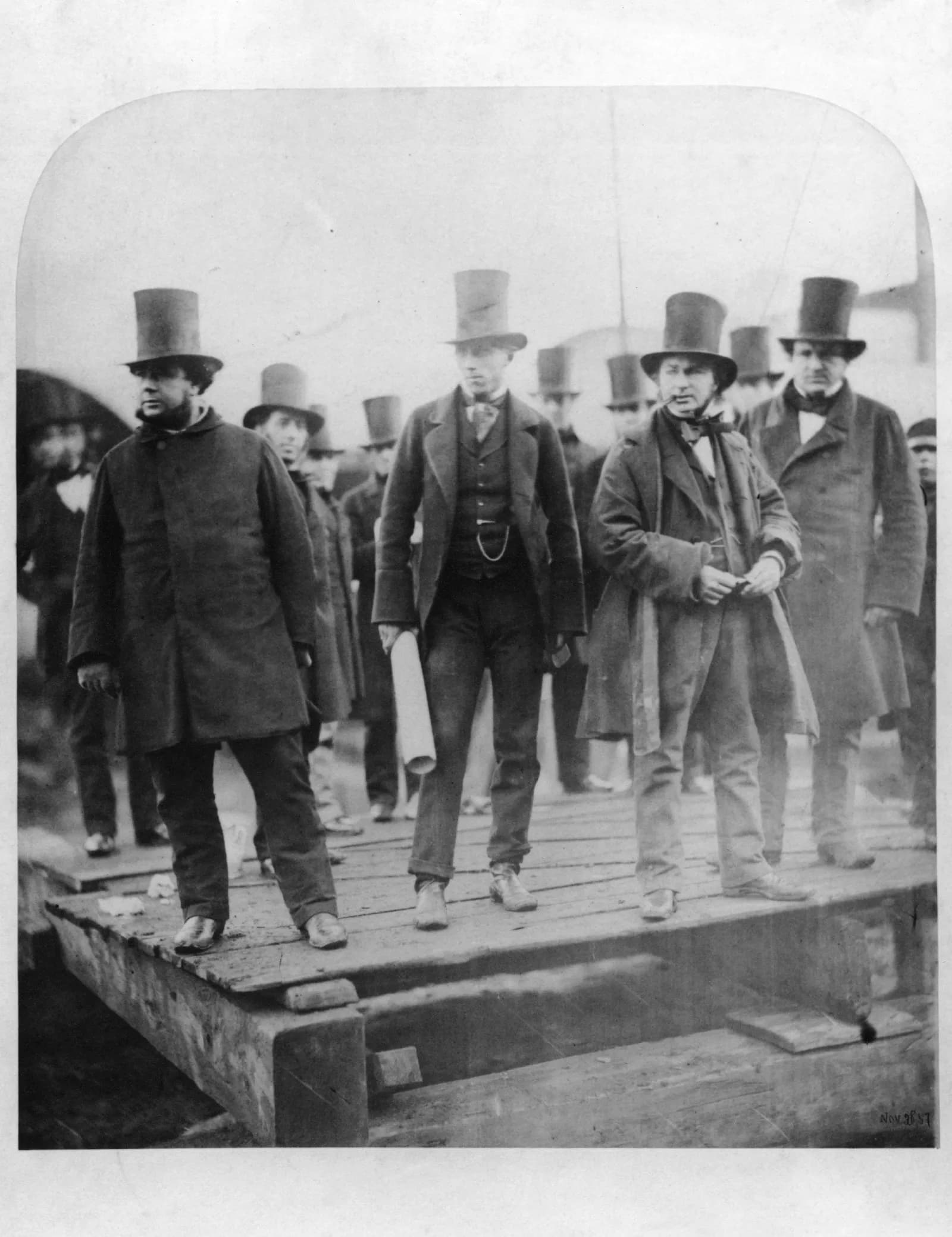 A black and white photograph showing a group of men wearing top hats and long coats standing on a wooden platform.