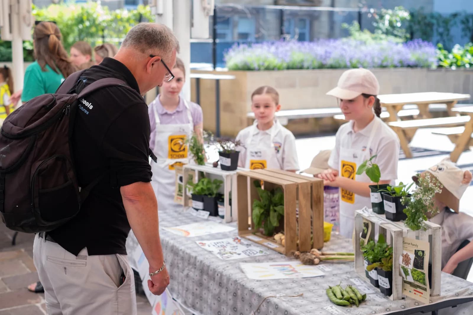 A man wearing a black shirt and backpack talks to three girls in aprons behind a table laden with potted plants and produce.
