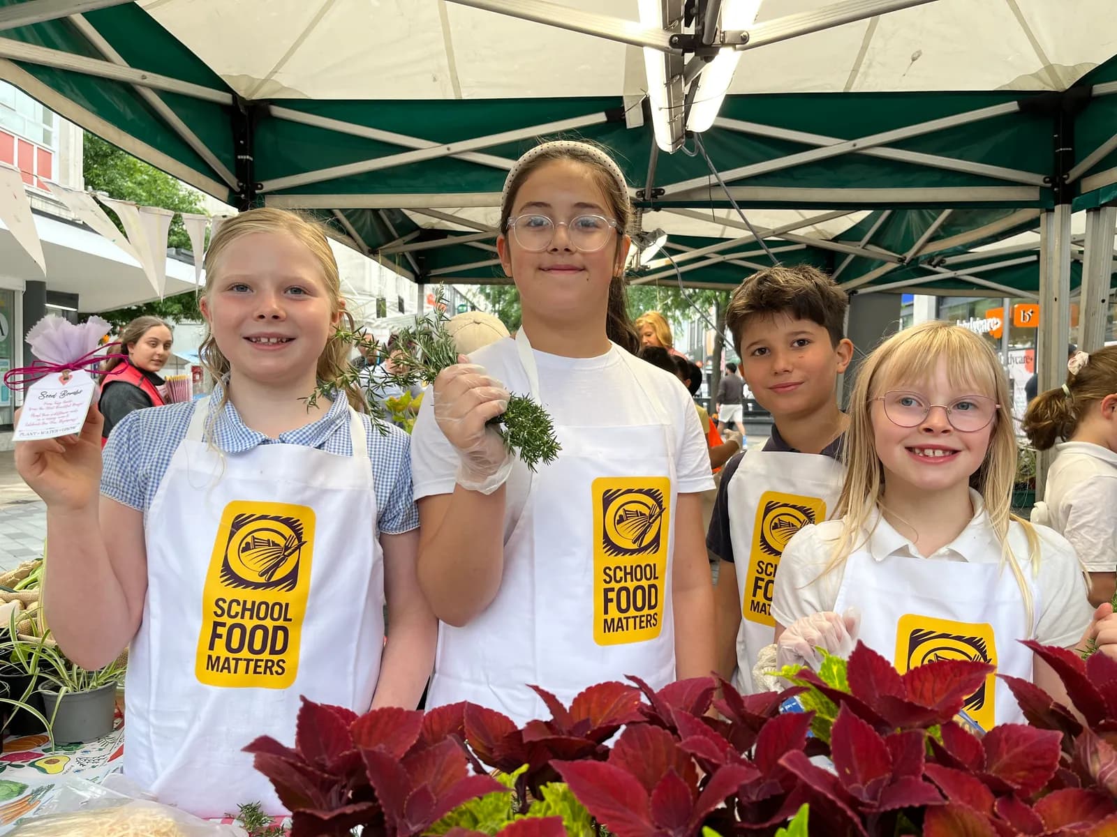 Four children smiling and wearing white aprons with the "School Food Matters" logo are standing behind a table with plants, with two holding items to sell.