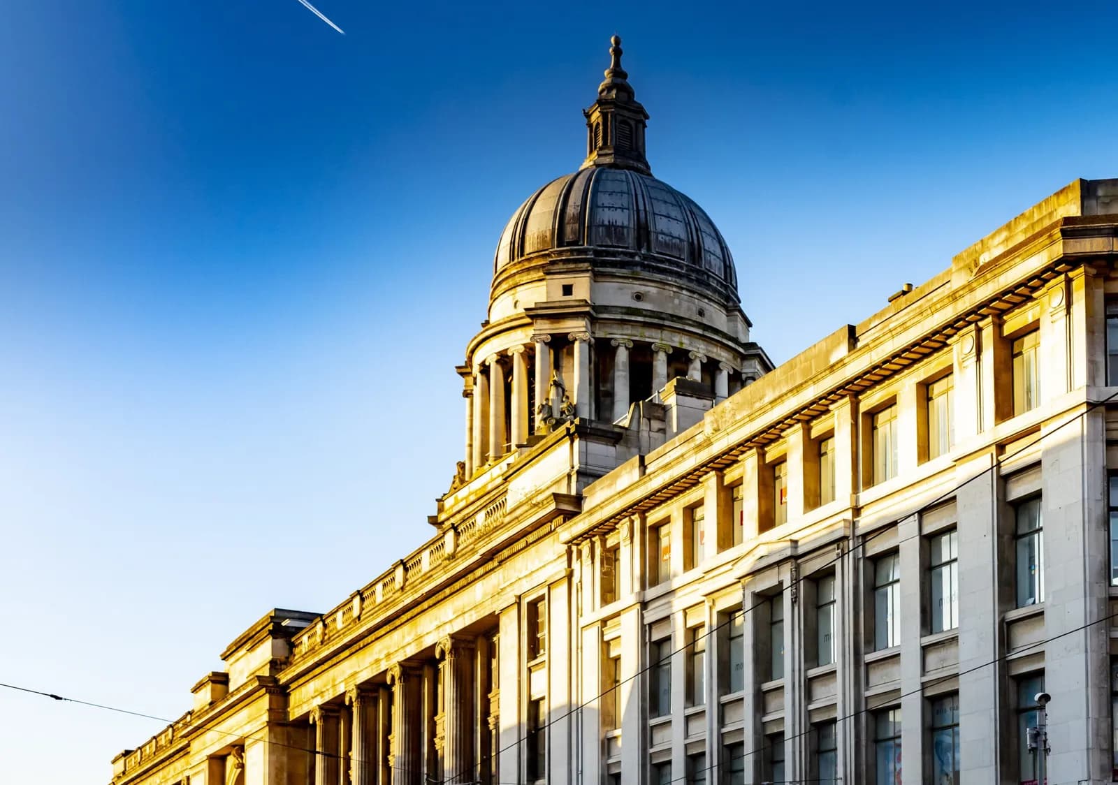 A grand building featuring a large domed roof and classical columns, with a modern building adjacent to it under a clear blue sky with a faint contrail.