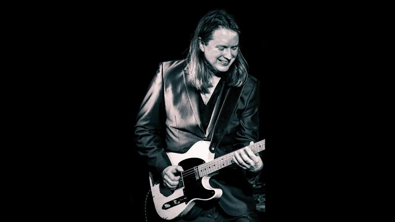 A man with long hair, wearing a dark suit, is shown from the waist up, smiling and playing an electric guitar against a black background.