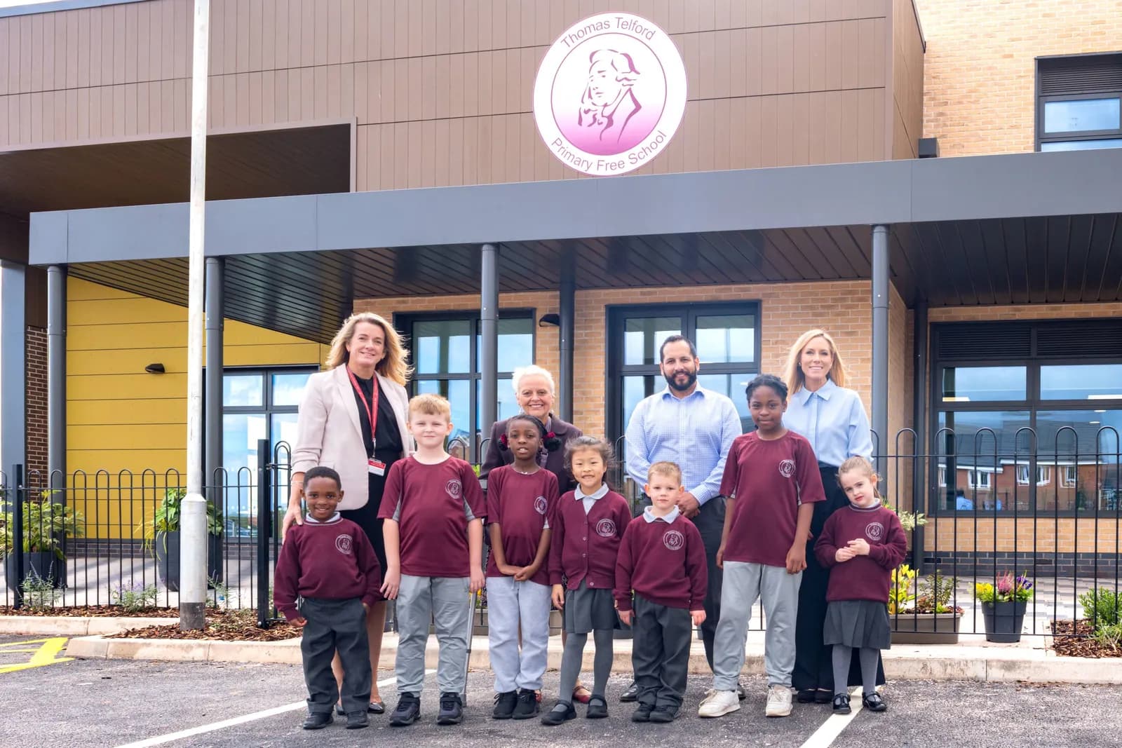 A group of adults and children in school uniform stand in front of the modern Thomas Telford Primary Free School building.