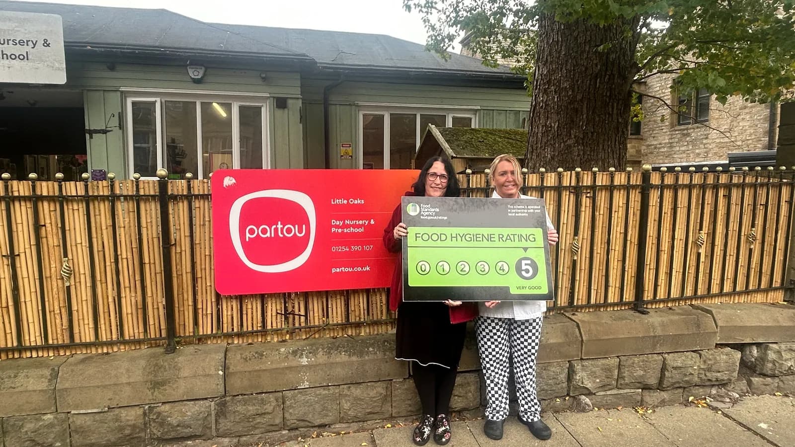 Two women stand in front of a bamboo fence and building, holding signs; one red with the word "partou" and information about a pre-school, and one black and green with a "Food Hygiene Rating 5, Very Good."